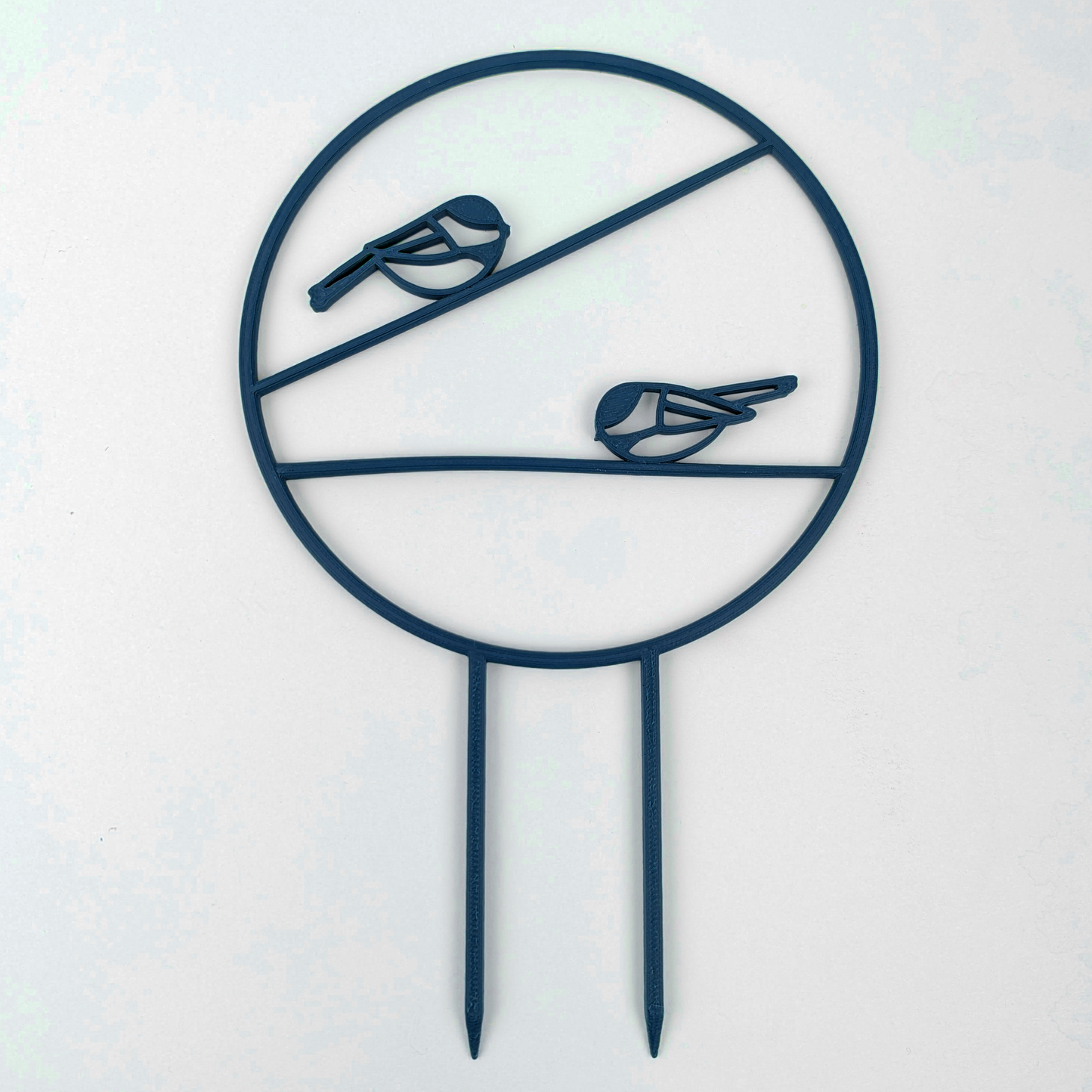 The chickadee trellis. A circle with two legs on the bottom for stakes. Across the middle are two branches running horizontally one on top of the other, each with a chickadee on it. Dark Blue colour against a white background.