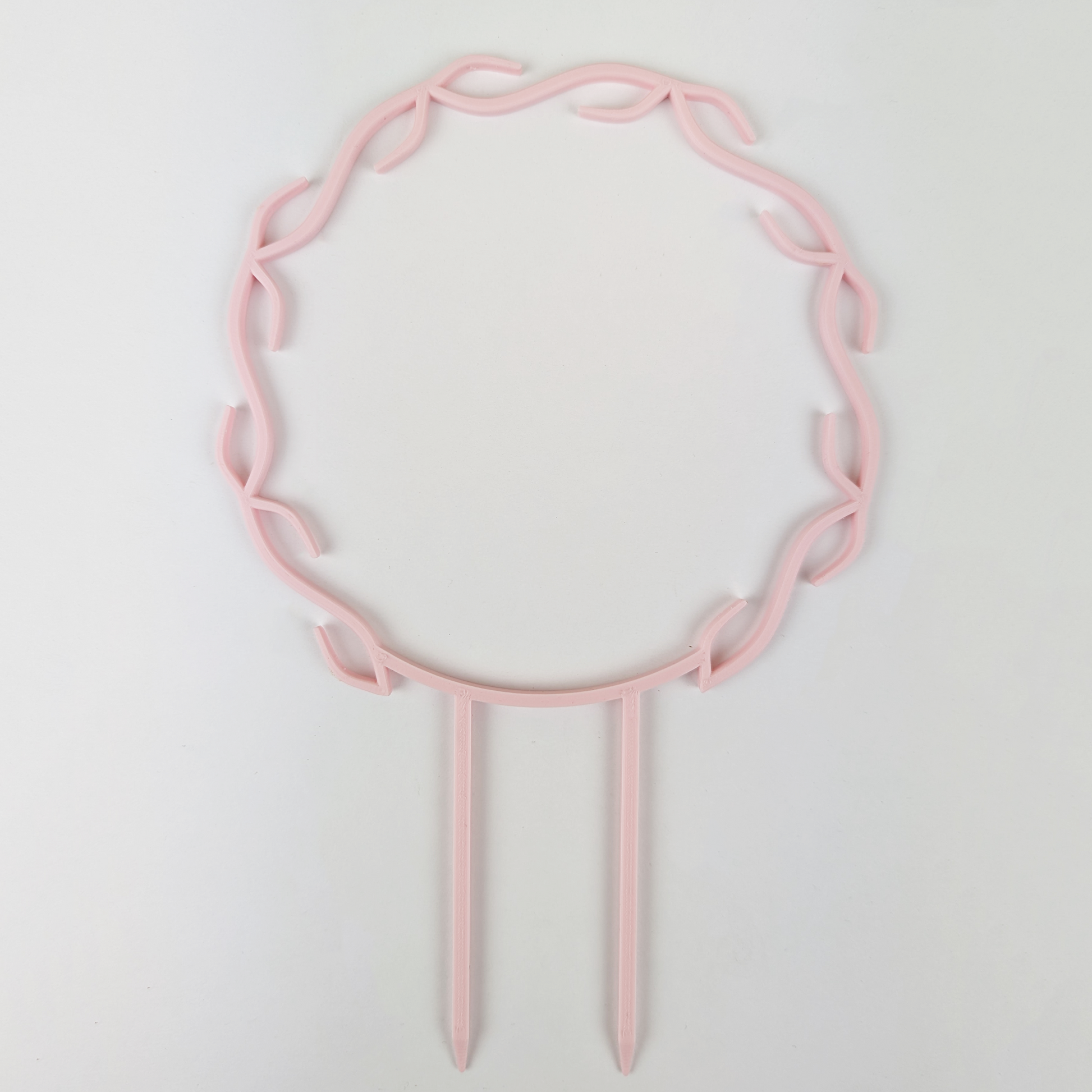 A wreath trellis. It has little hooks that go around in a circle, almost looking like a wreath. It is in pastel pink colour against a white background.